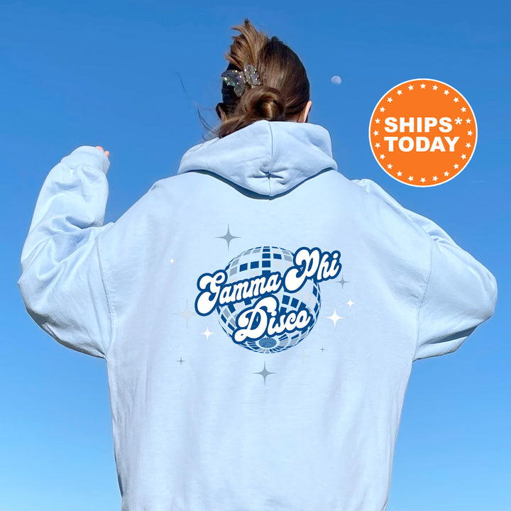 A person wearing a light blue hoodie with a graphic design on the back, including a disco ball and stars, and a circular orange sign with the text "SHIPS TODAY".