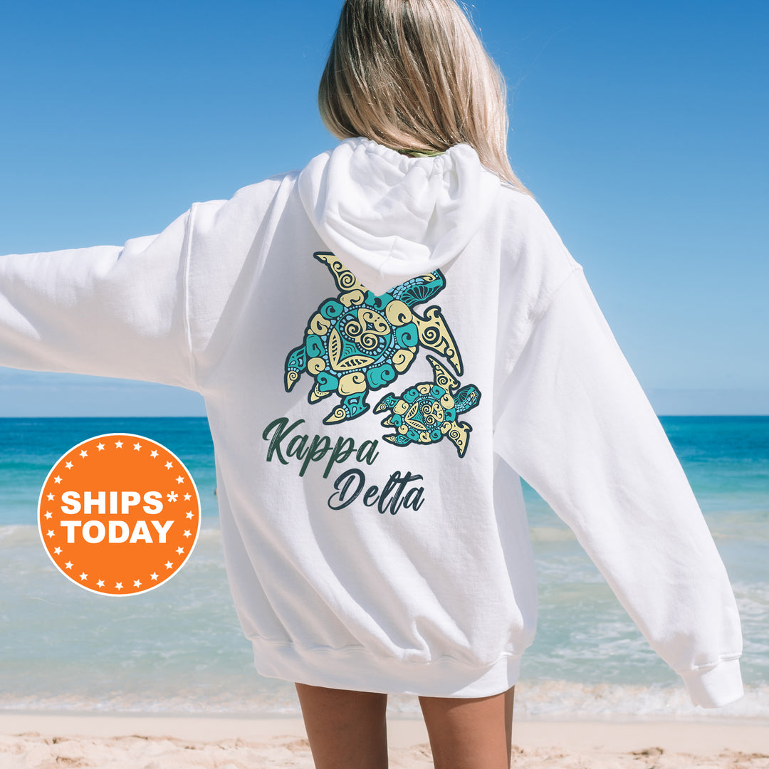 A woman wearing a white hoodie with a colorful graphic of a turtle on the back, standing on a beach with the ocean in the background.