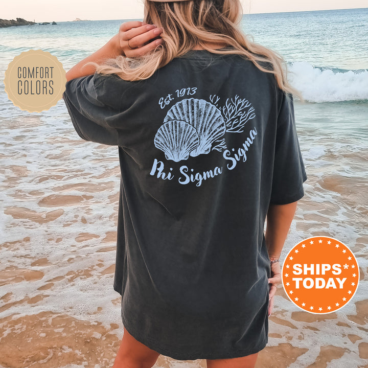 A woman wearing a black t-shirt with a graphic design of seashells and the text "Phi Sigma Sigma" standing on a beach.