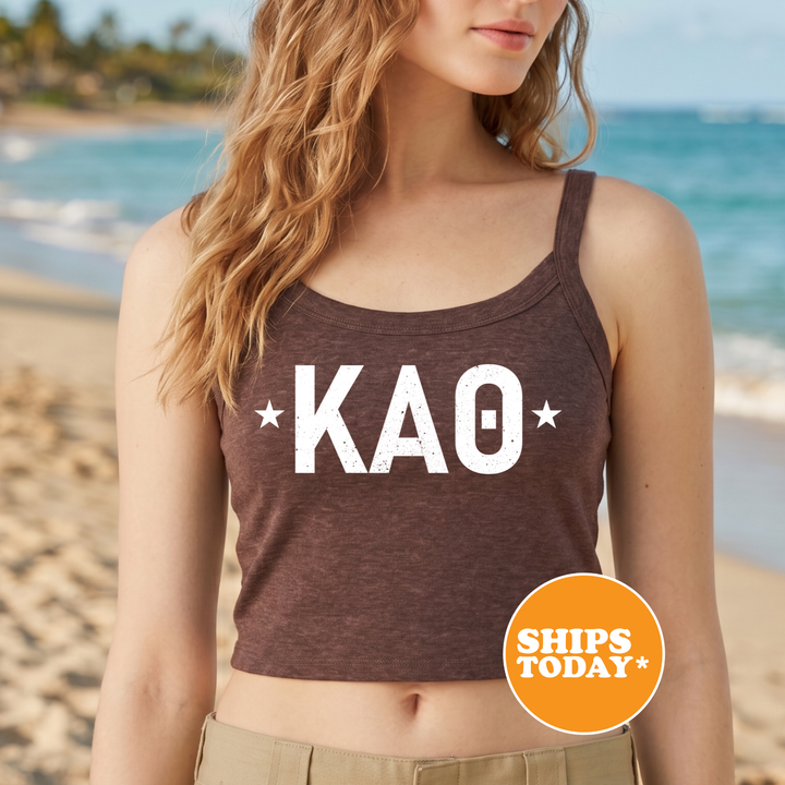 A woman wearing a brown crop top with the letters "KAO" printed on it, standing on a beach with the ocean in the background.