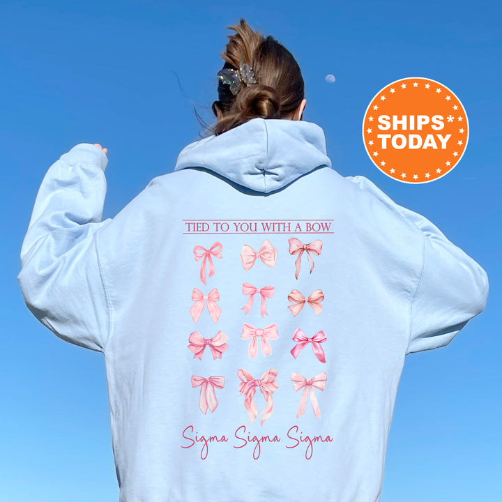 A person wearing a light blue hoodie with a graphic of pink bows on the back, standing against a clear blue sky.