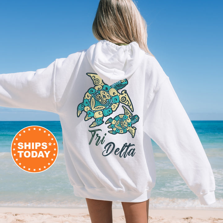 A woman wearing a white hoodie with a colorful turtle design on the back, standing on a beach with the ocean in the background.