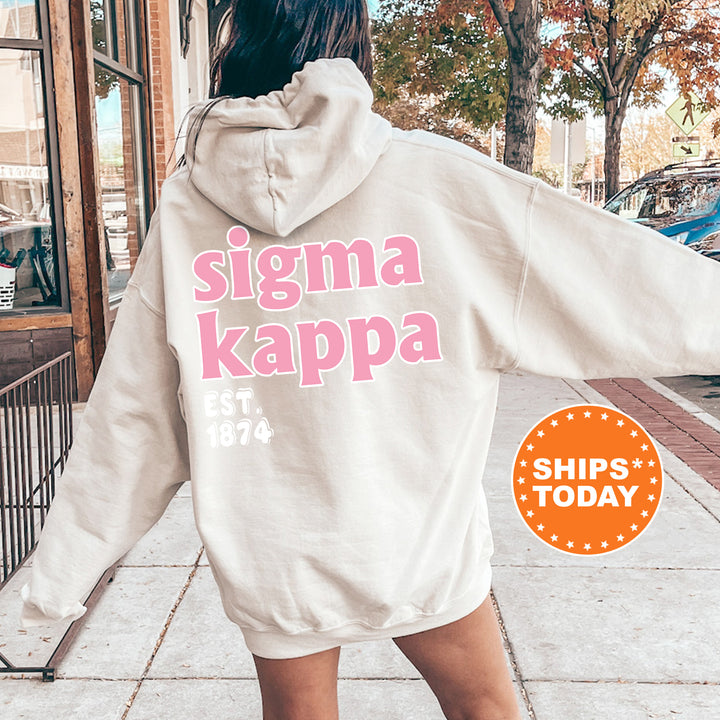 A woman wearing a white hoodie with pink text and logos, standing on a sidewalk.