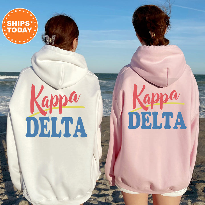 Two women wearing matching white and pink hoodies with the text "Kappa Delta" printed on them, standing on a beach with the ocean in the background.