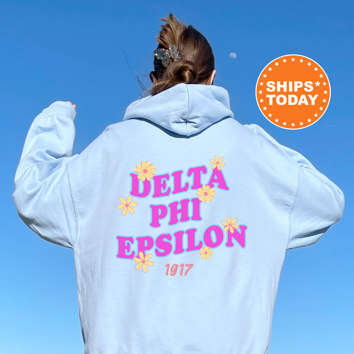 A person wearing a white hoodie with the Delta Phi Epsilon logo and the year 1977 printed on it, standing against a clear blue sky.