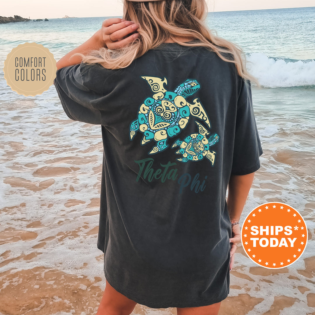 A woman wearing a black t-shirt with a colorful turtle design on the back, standing on a beach with the ocean in the background.