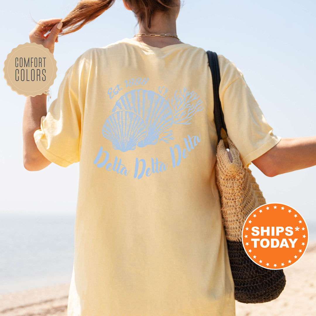 A woman wearing a yellow Delta Delta Delta t-shirt with a seashell design, standing on a beach with her hair in a ponytail.