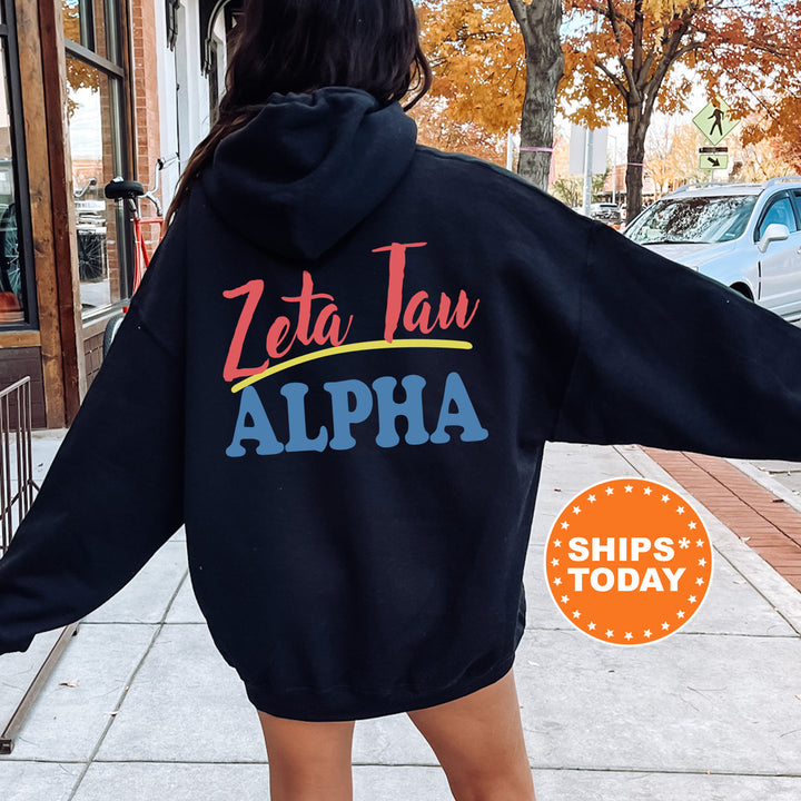 A person wearing a black hoodie with the text "Zeta Tan Alpha" printed on it, standing on a sidewalk with a car and a building in the background.