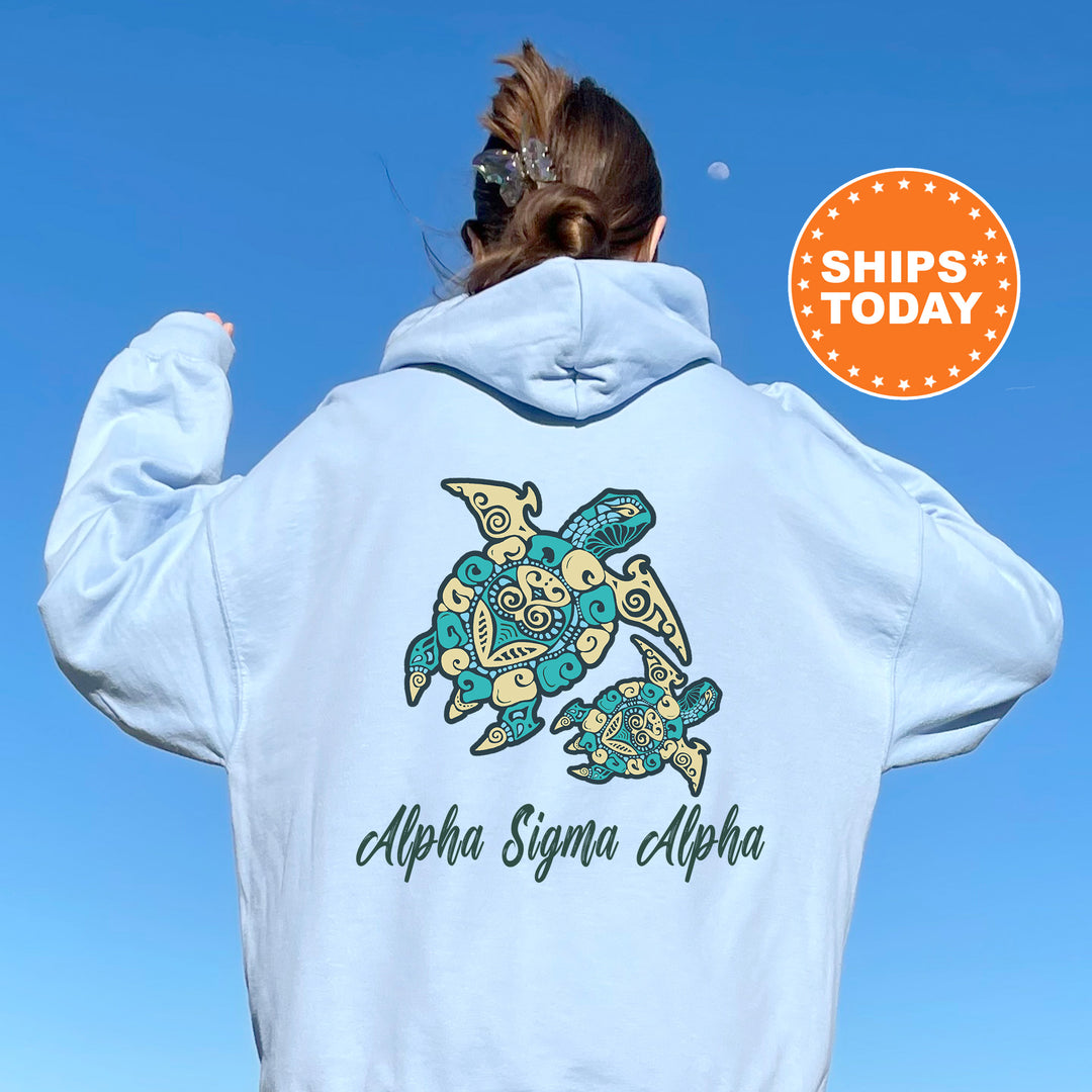 A person wearing a light blue hoodie with a graphic of a turtle and the text "Alpha Sigma Alpha" printed on it, standing against a clear blue sky.