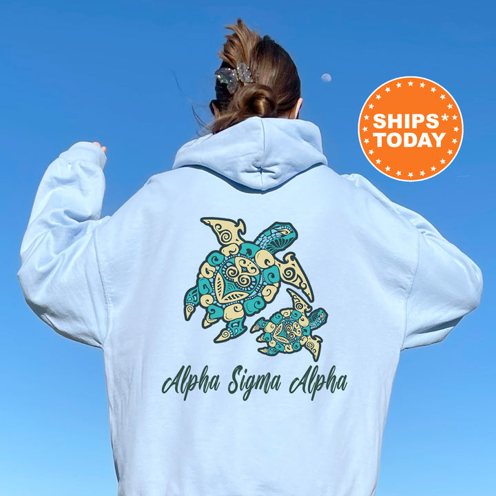 A person wearing a light blue hoodie with a graphic of a turtle and the text "Alpha Sigma Alpha" printed on it, standing against a clear blue sky.