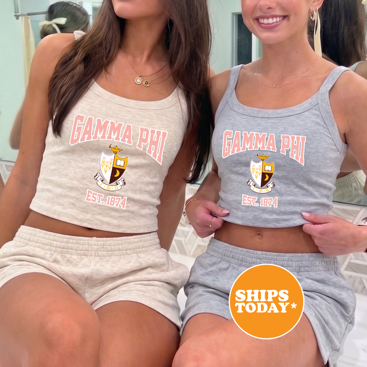 Two young women wearing matching tank tops with the text "GAMA PHI" and "EST. 1874" printed on them, posing together in front of a mirror.