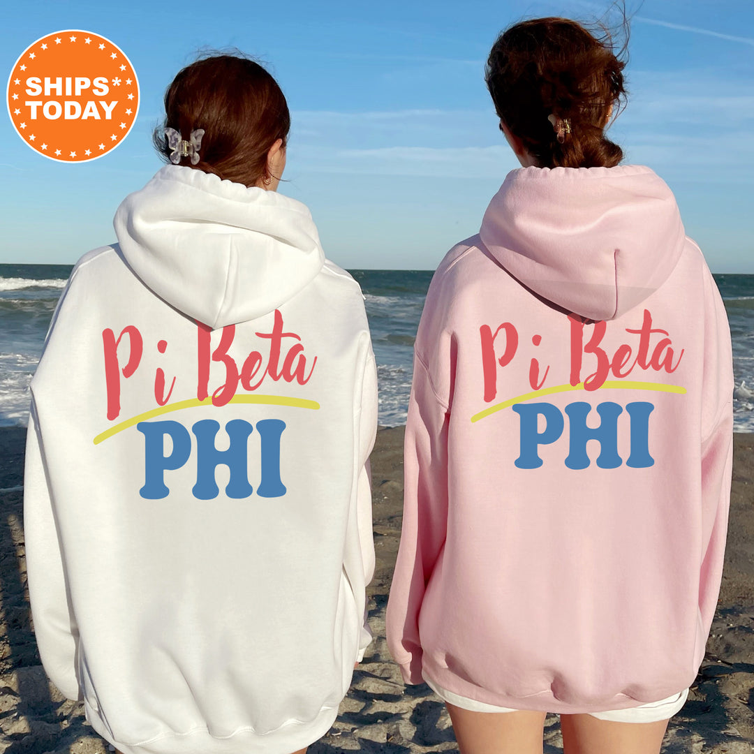 Two women wearing hooded sweatshirts with the text "Pi Beta PHI" printed on them, standing on a beach with the ocean in the background.