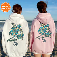 Two women wearing matching hoodies with a turtle design and text on the back, standing on a beach with the ocean in the background.