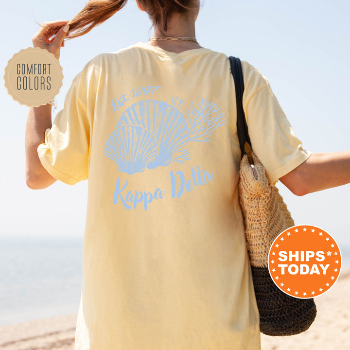 A woman wearing a yellow t-shirt with a blue design and text, standing on a beach with her hair in a ponytail.