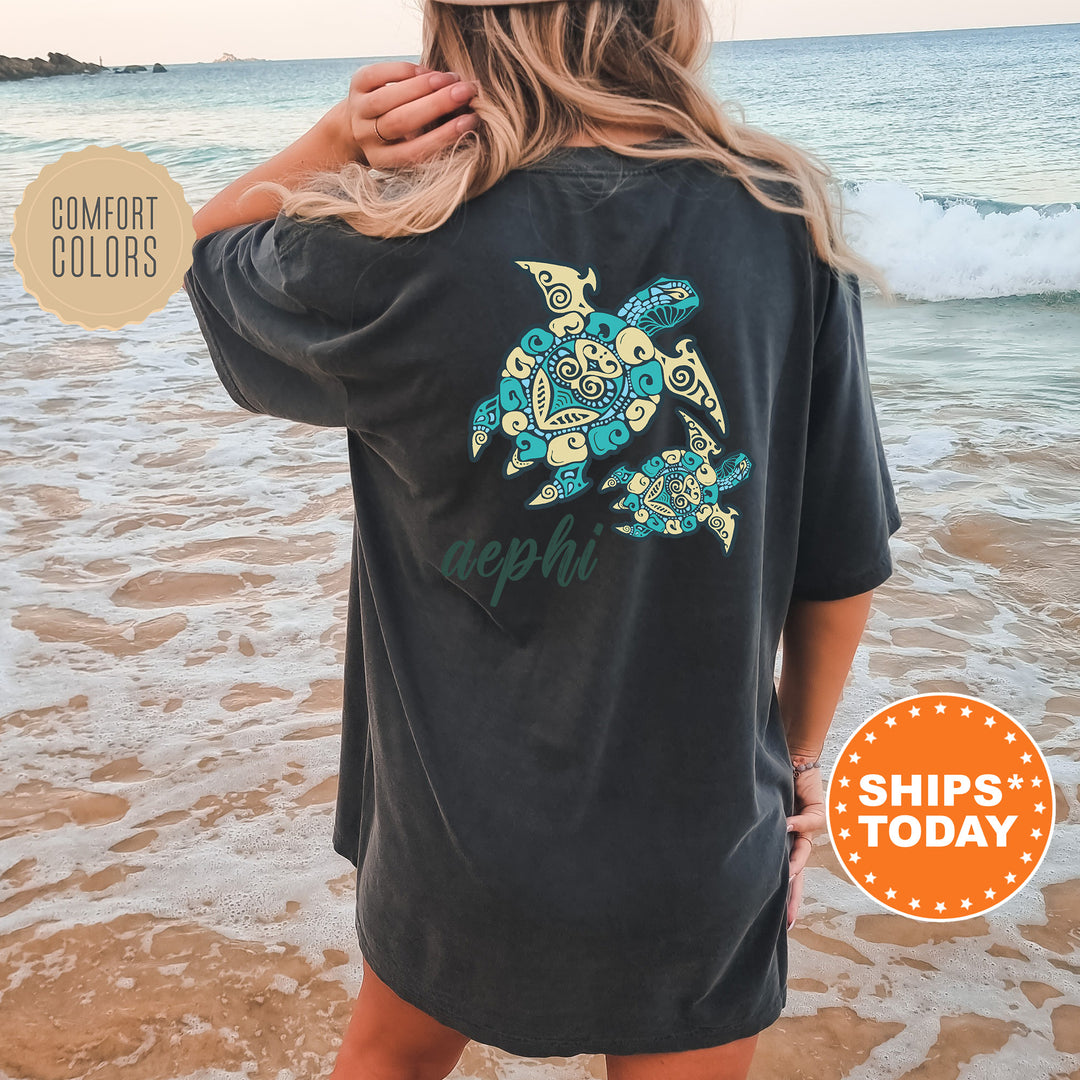 A woman wearing a black t-shirt with a colorful turtle design stands on a beach, with the ocean visible in the background.