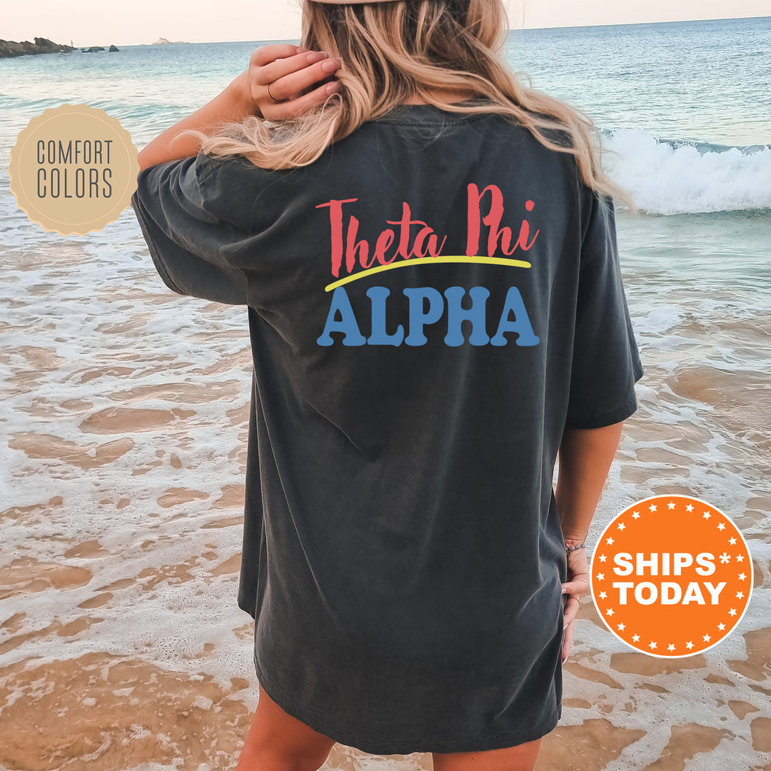 A woman wearing a black t-shirt with the text "Theta Phi Alpha" printed on it, standing on a beach with the ocean in the background.