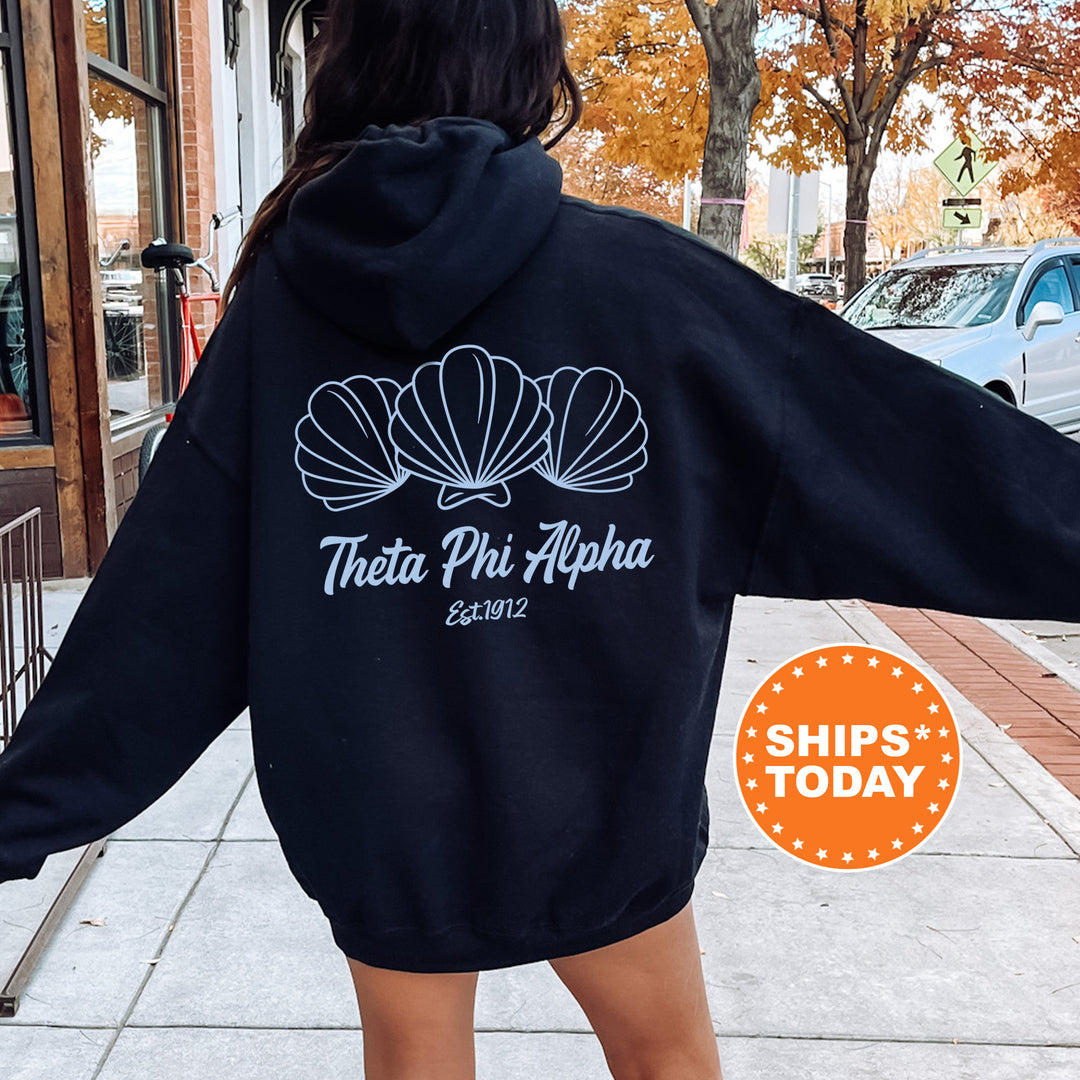 A person wearing a black hoodie with a white design of three shells and the text "Theta Phi Alpha Est. 1982" printed on it, standing on a sidewalk.