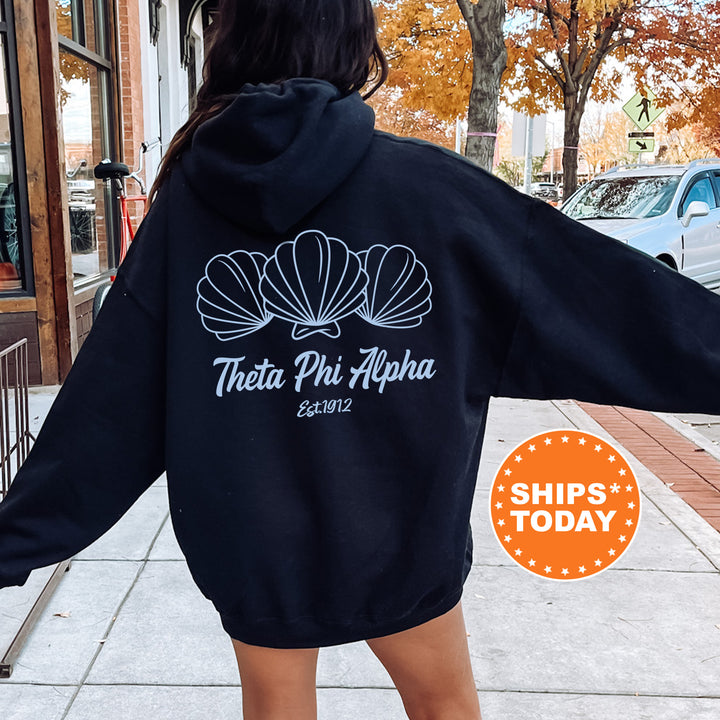 A person wearing a black hoodie with a white design of three shells and the text "Theta Phi Alpha Est. 1982" printed on it, standing on a sidewalk.
