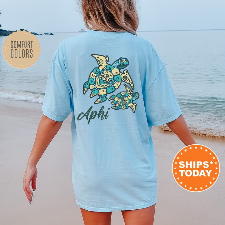 A woman wearing a light blue t-shirt with a graphic of a turtle on the back, standing on a beach with the ocean in the background.