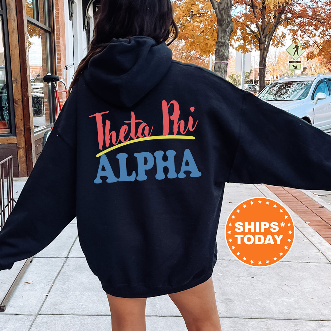 A person wearing a black hoodie with the text "Theta Phi Alpha" printed on it, standing on a sidewalk with a car and a building in the background.