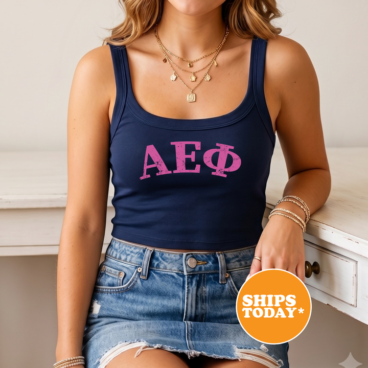 A woman wearing a navy blue crop top with the letters "AE Φ π" printed on it, paired with denim shorts and bracelets. She is sitting on a white surface, and there is a circular orange sign with white text that reads "SHIPS TODAY".