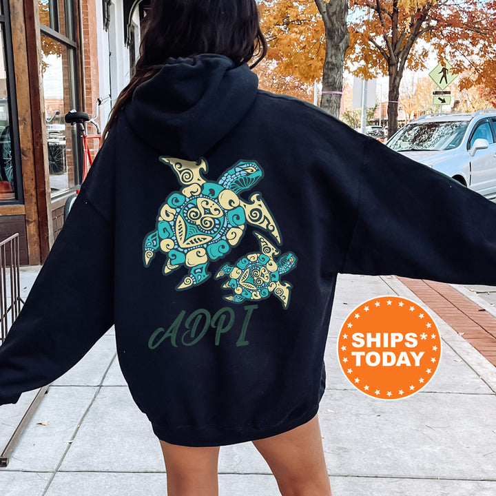 A person wearing a black hoodie with a colorful turtle design on the back, standing on a sidewalk with a car and a building in the background.