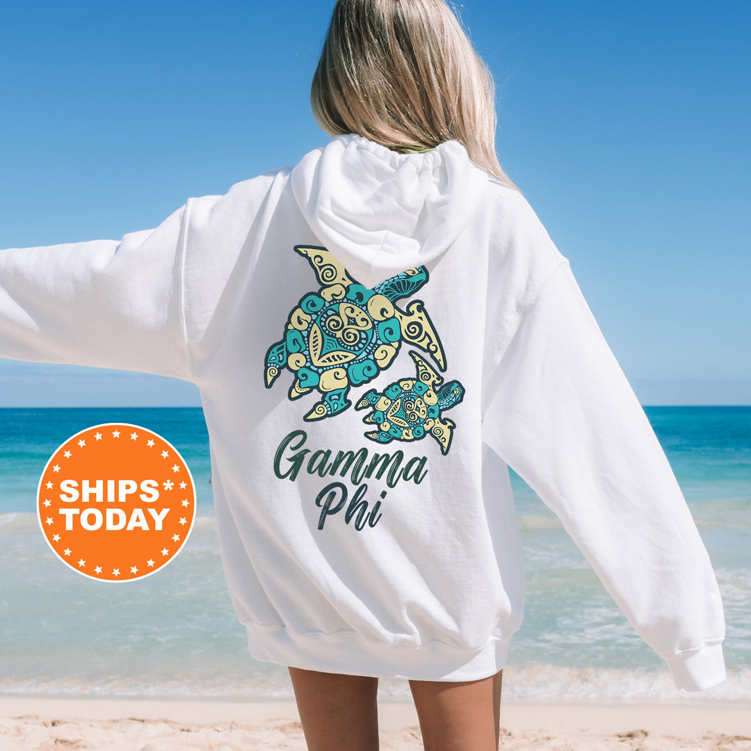 A woman wearing a white hoodie with a graphic of a turtle on the back, standing on a beach with the ocean in the background.