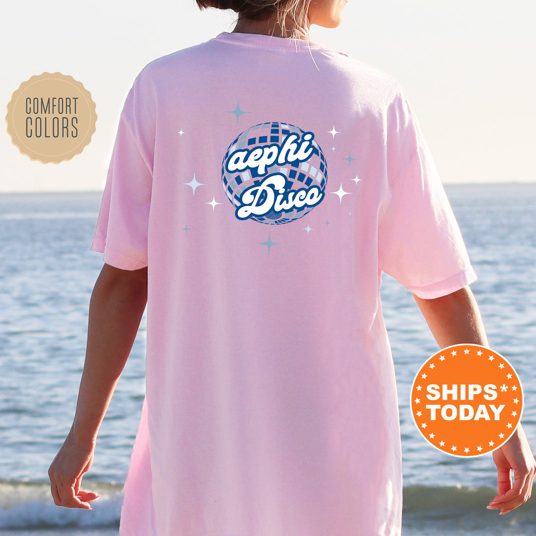 A person wearing a pink t-shirt with a graphic design of a disco ball and stars, standing near the ocean.