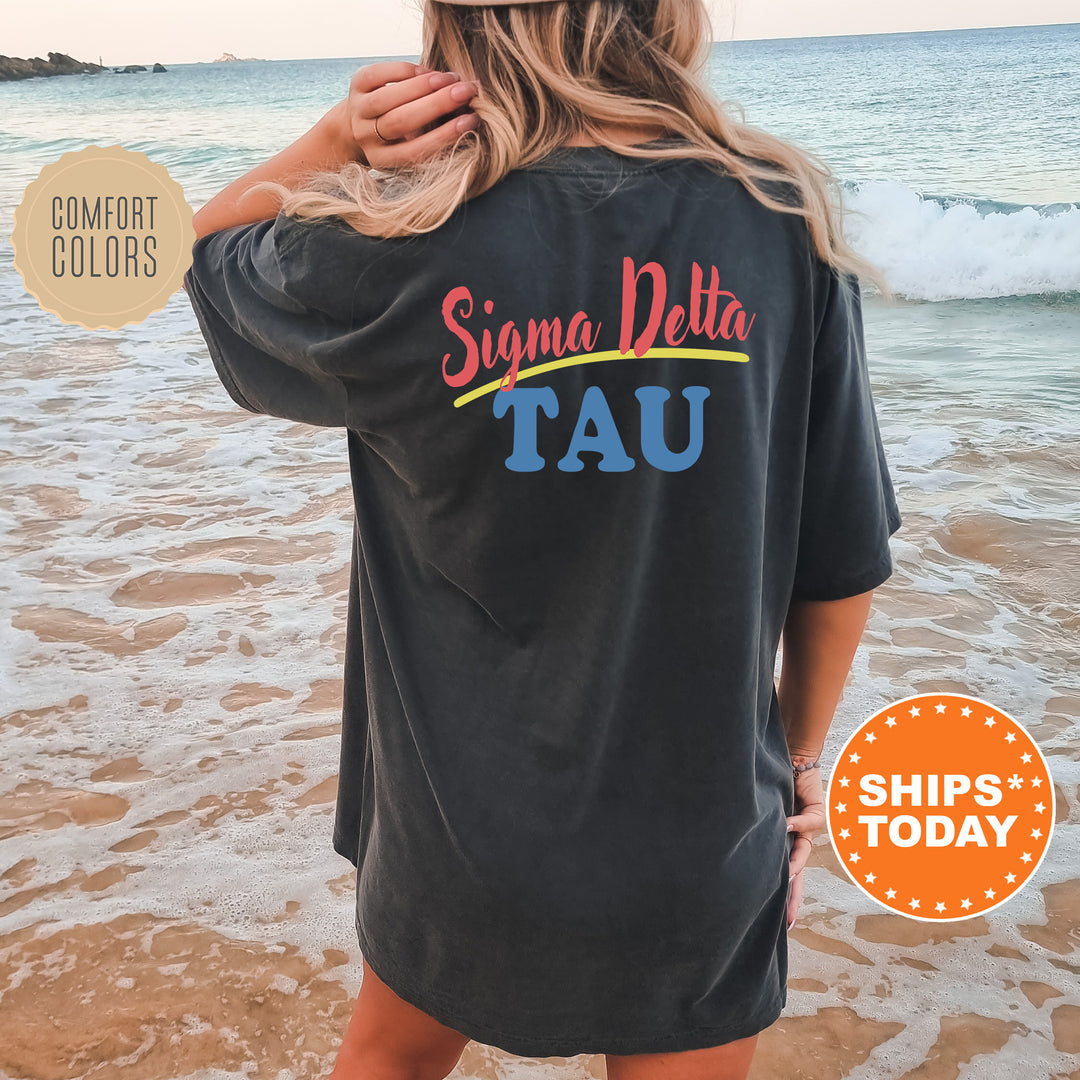A woman wearing a black t-shirt with the text "Signa Delta Tau" printed on it, standing on a beach with the ocean in the background.