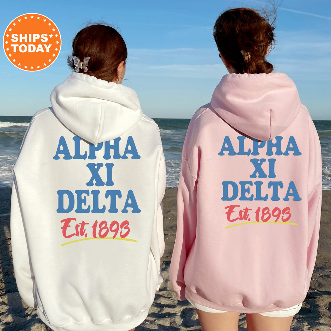 Two women wearing matching white and pink hoodies with the text "ALPA XI DELTA Est. 1893" printed on them, standing on a beach with the ocean in the background.