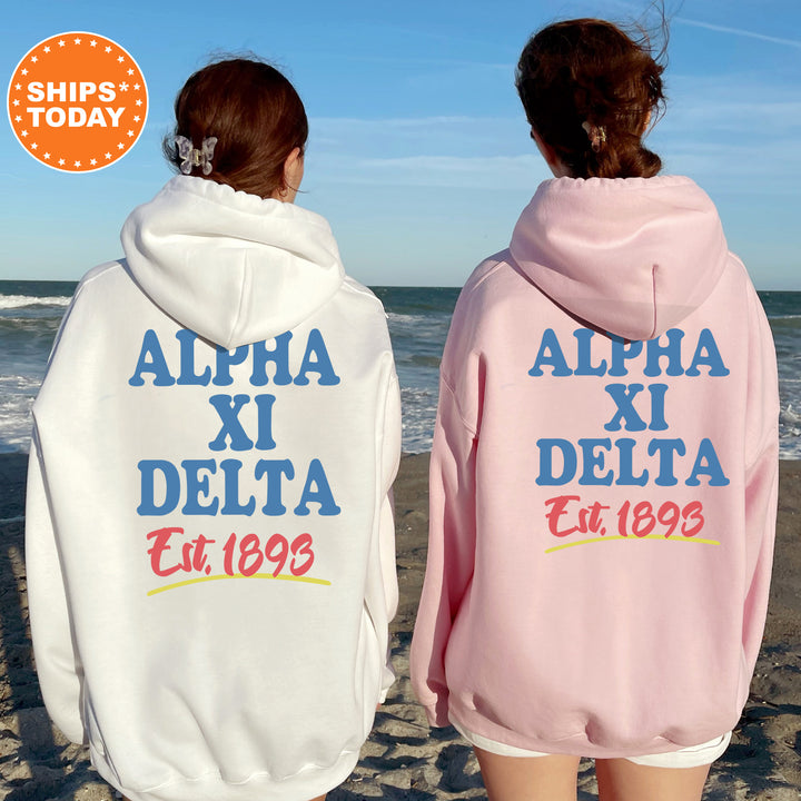 Two women wearing matching white and pink hoodies with the text "ALPA XI DELTA Est. 1893" printed on them, standing on a beach with the ocean in the background.