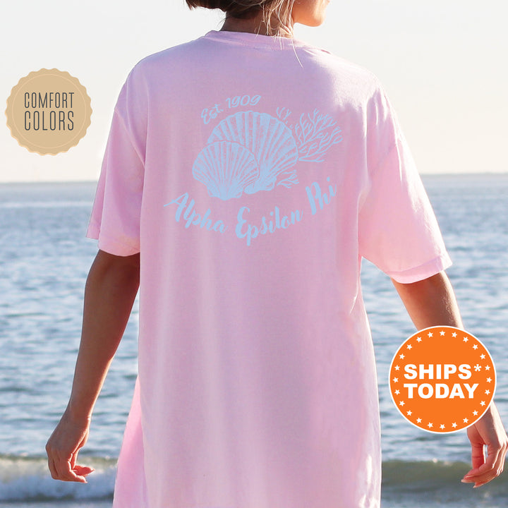 A person wearing a pink t-shirt with a graphic design of a shell and the text "Aloha Epilon Phi" is standing on a beach with the ocean in the background.