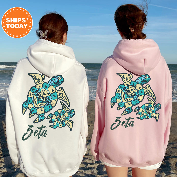 Two women standing on a beach, wearing matching white and pink hoodies with a turtle design and the name 'Zeta' printed on them.
