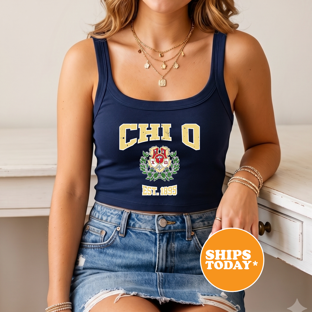 A woman wearing a navy blue crop top with the letters "CHI Q" printed on it, a denim skirt, and multiple necklaces. She is sitting on a white surface with a sign that says "SHIPS TODAY".