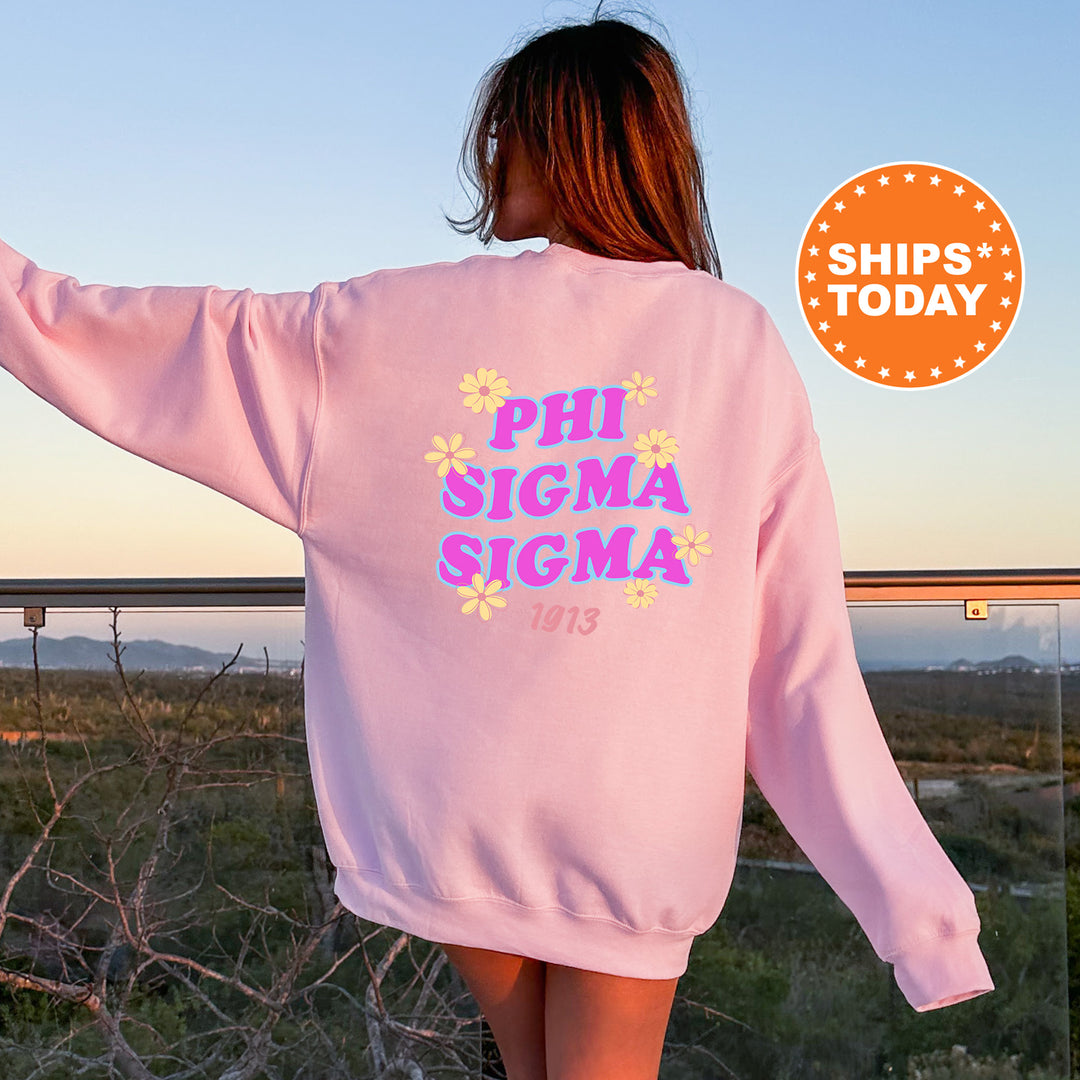 A woman wearing a pink sweatshirt with the text "PHI SIGMA SIGMA" printed on it, standing in front of a scenic outdoor landscape.
