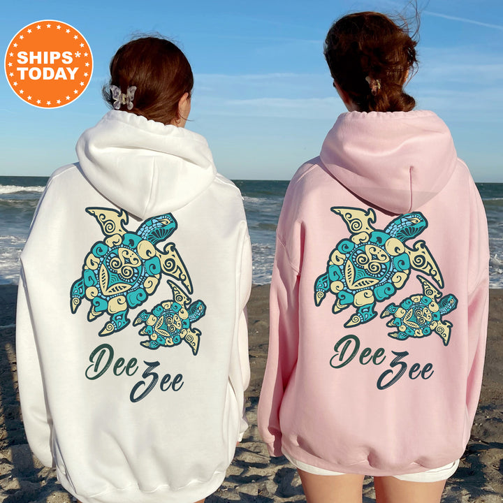 Two women wearing matching hoodies with a turtle design on the back, standing on a beach with the ocean in the background.