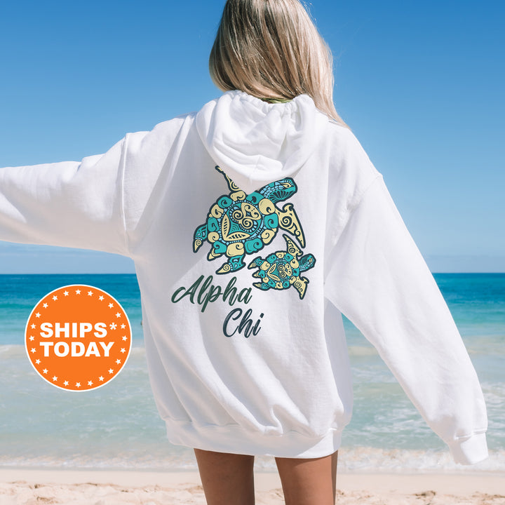 A woman wearing a white hoodie with a graphic of a turtle and the text "Alpha Chi" printed on it, standing on a beach with the ocean in the background.