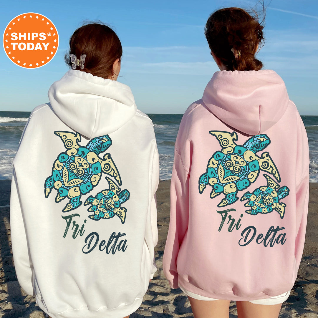 Two women wearing matching Tri Delta hoodies stand on a beach, facing the ocean.