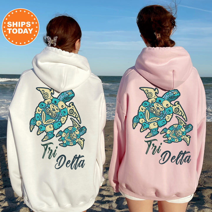 Two women wearing matching Tri Delta hoodies stand on a beach, facing the ocean.