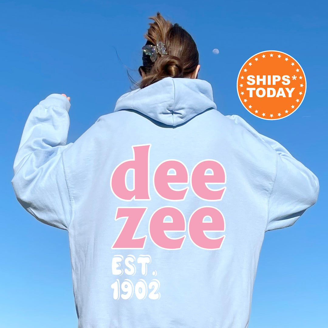A person wearing a light blue hoodie with the text "Dee Zee" and "EST. 1902" printed on it, standing against a clear blue sky.