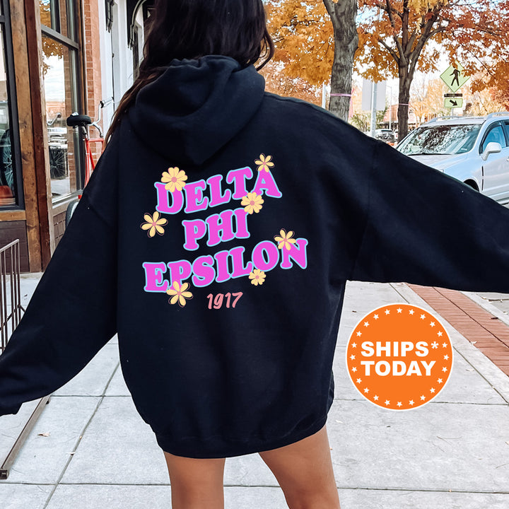 A person wearing a black hoodie with the Delta Phi Epson logo and the year 1977 printed on it, standing on a sidewalk with a car and a tree in the background.