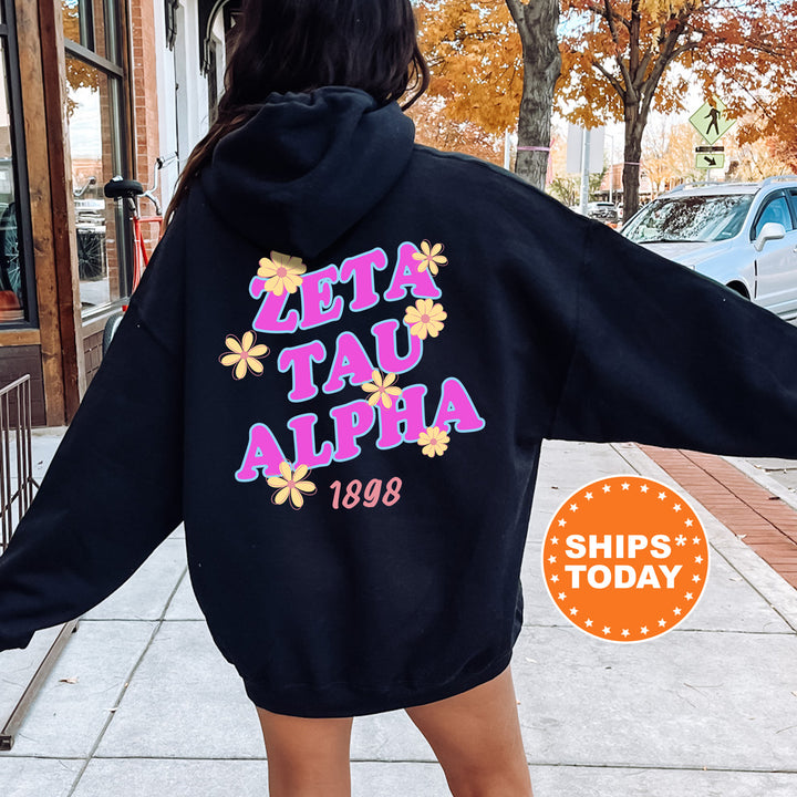A person wearing a black hoodie with the text "ZETA TAU ALPHA 1898" printed on it, standing on a sidewalk with a car and a tree in the background.