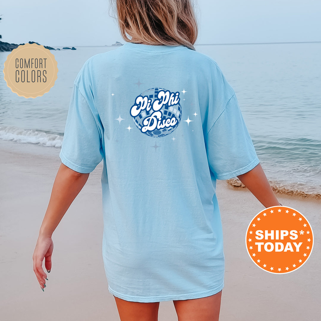 A person wearing a light blue t-shirt with a graphic design on the back is standing on a beach, with the ocean visible in the background.