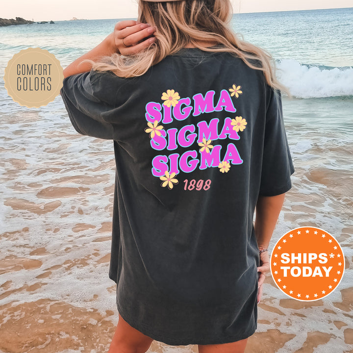 A woman wearing a black t-shirt with the text "SIGMA SIGMA SIGMA" printed on it, standing on a beach with the ocean in the background.
