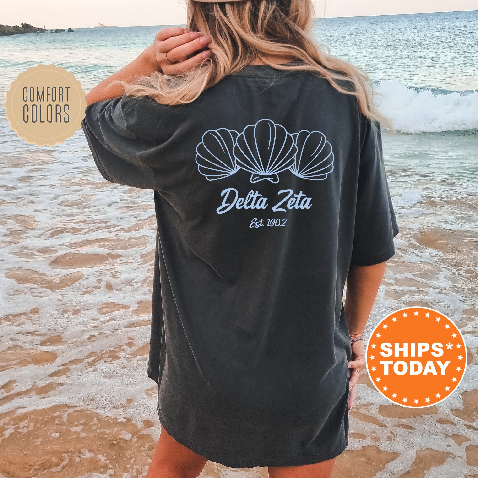 A woman wearing a black t-shirt with a graphic design of three shells on the back, standing on a beach with the ocean in the background.