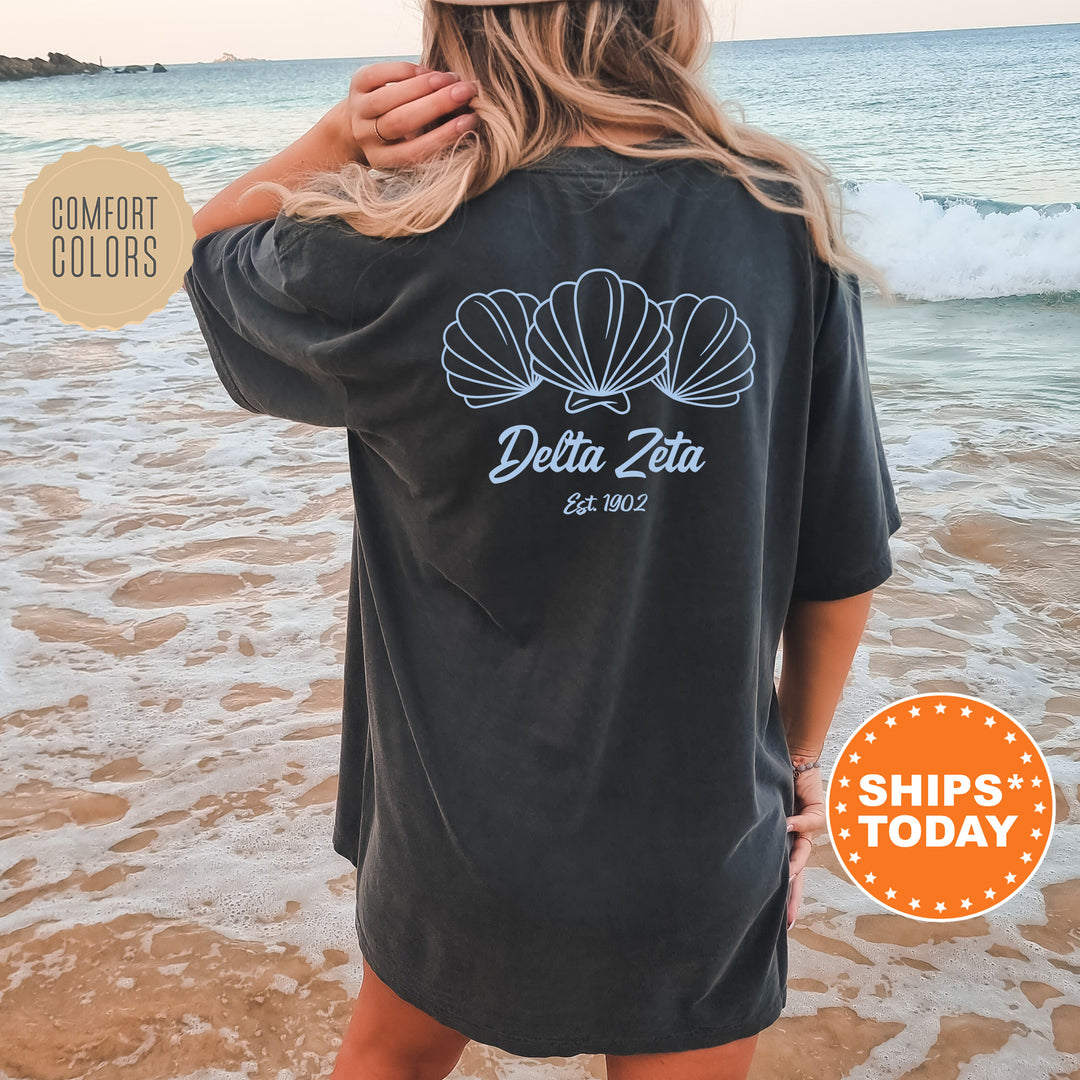 A woman wearing a black t-shirt with a graphic design of three shells on the back, standing on a beach with the ocean in the background.