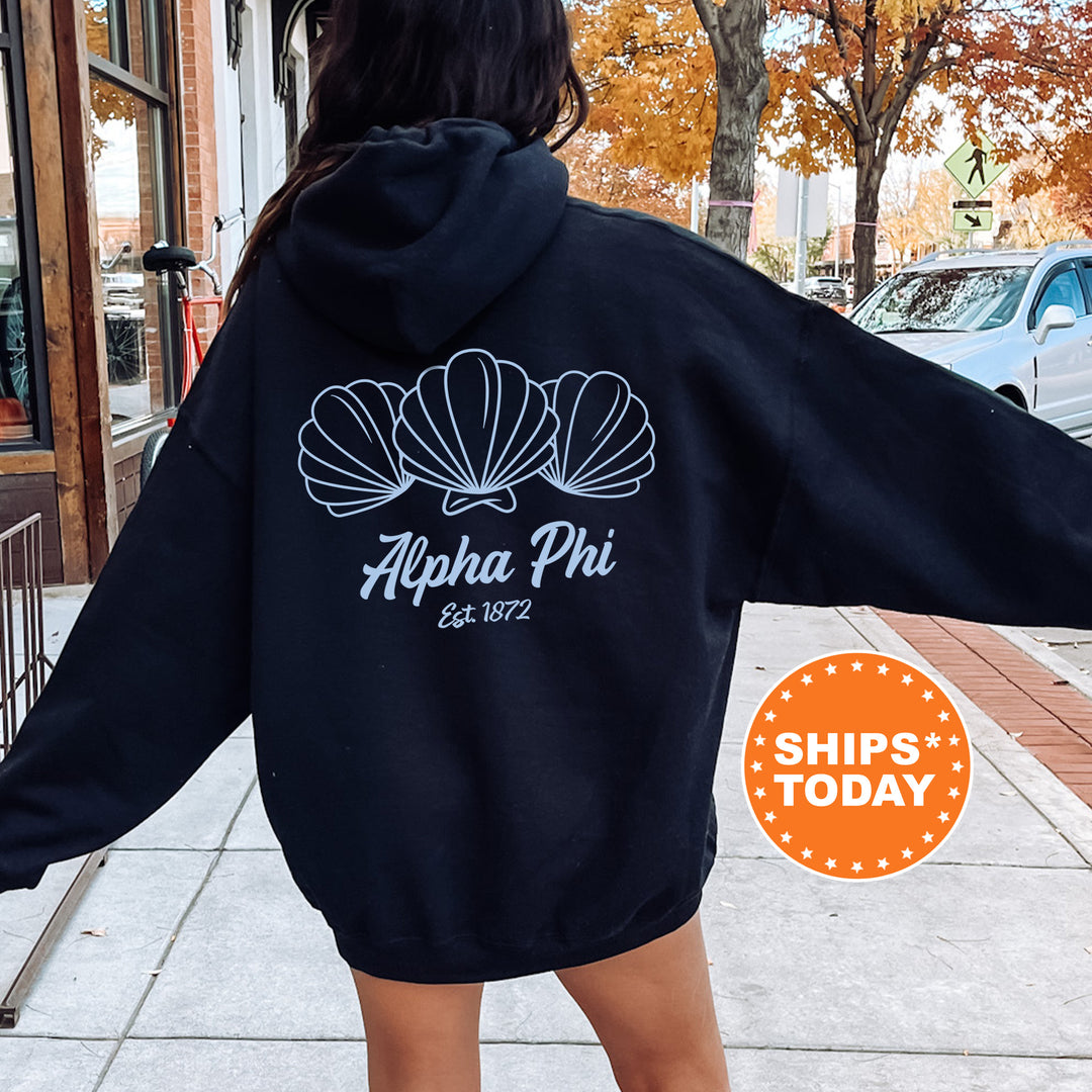 A person wearing a black hoodie with a graphic design of three shells and the text "Alpha Phi Est. 1872" printed on it, standing on a sidewalk.