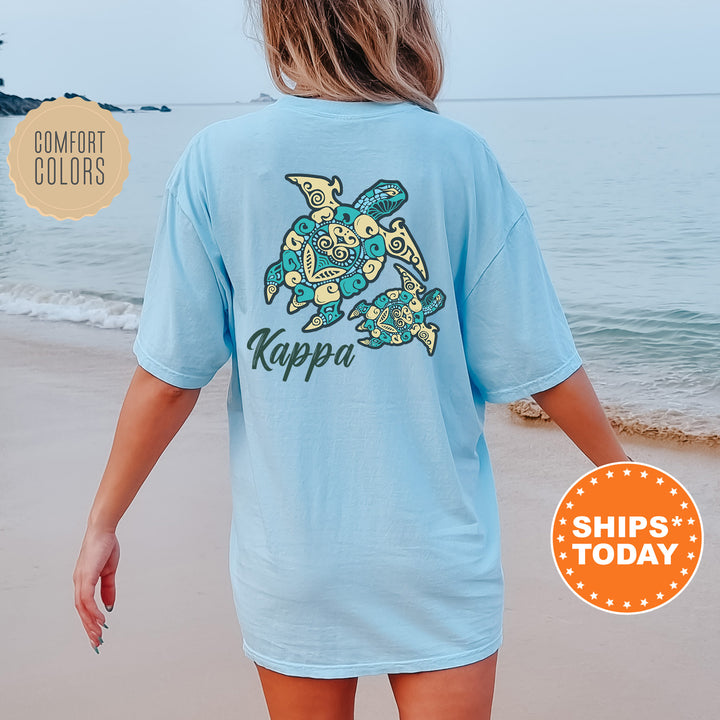 A woman wearing a light blue t-shirt with a colorful turtle design on the back, standing on a beach with the ocean in the background.