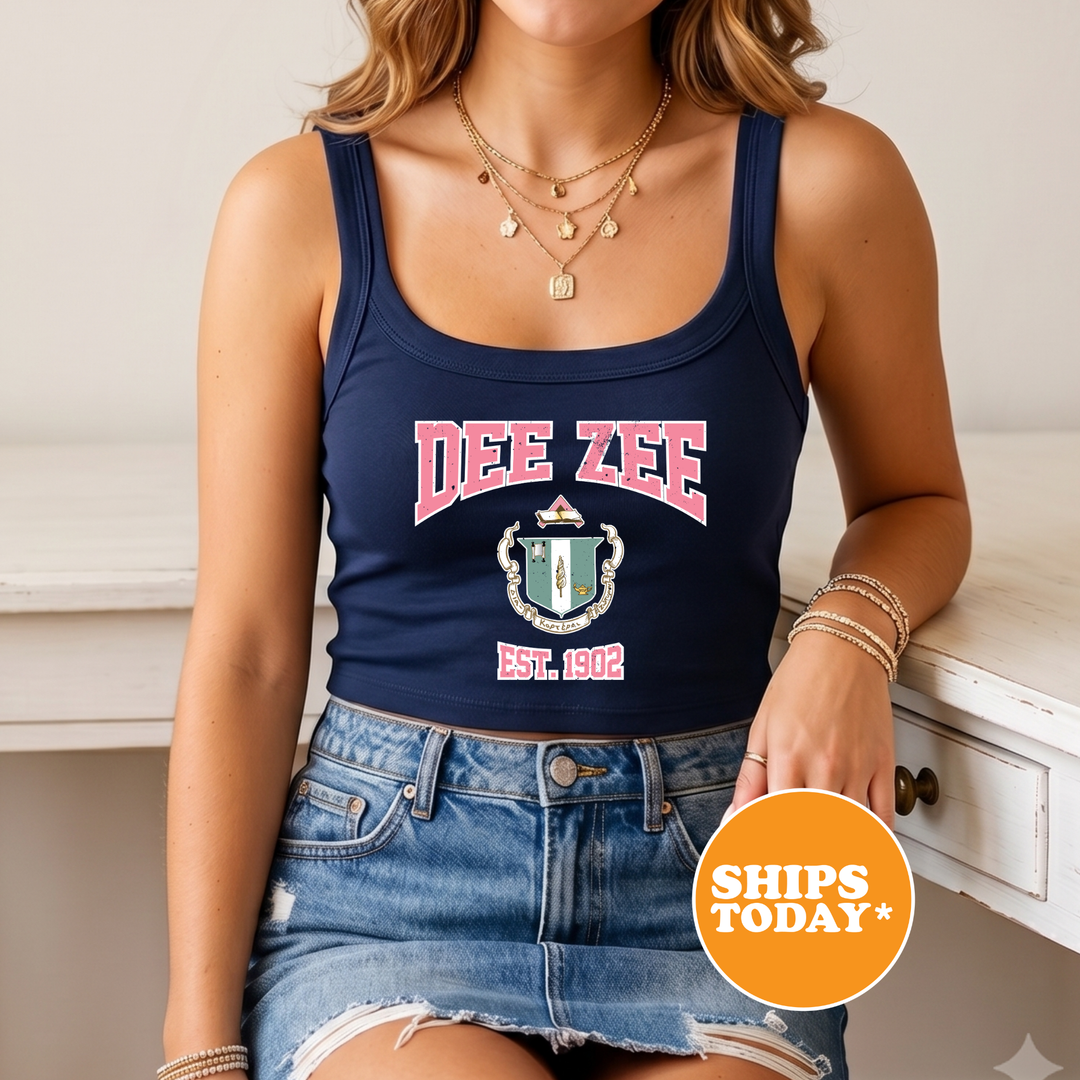 A woman wearing a navy blue crop top with the text "Dee Zee EST 1902" printed on it, paired with denim shorts. She is also wearing multiple necklaces and bracelets.