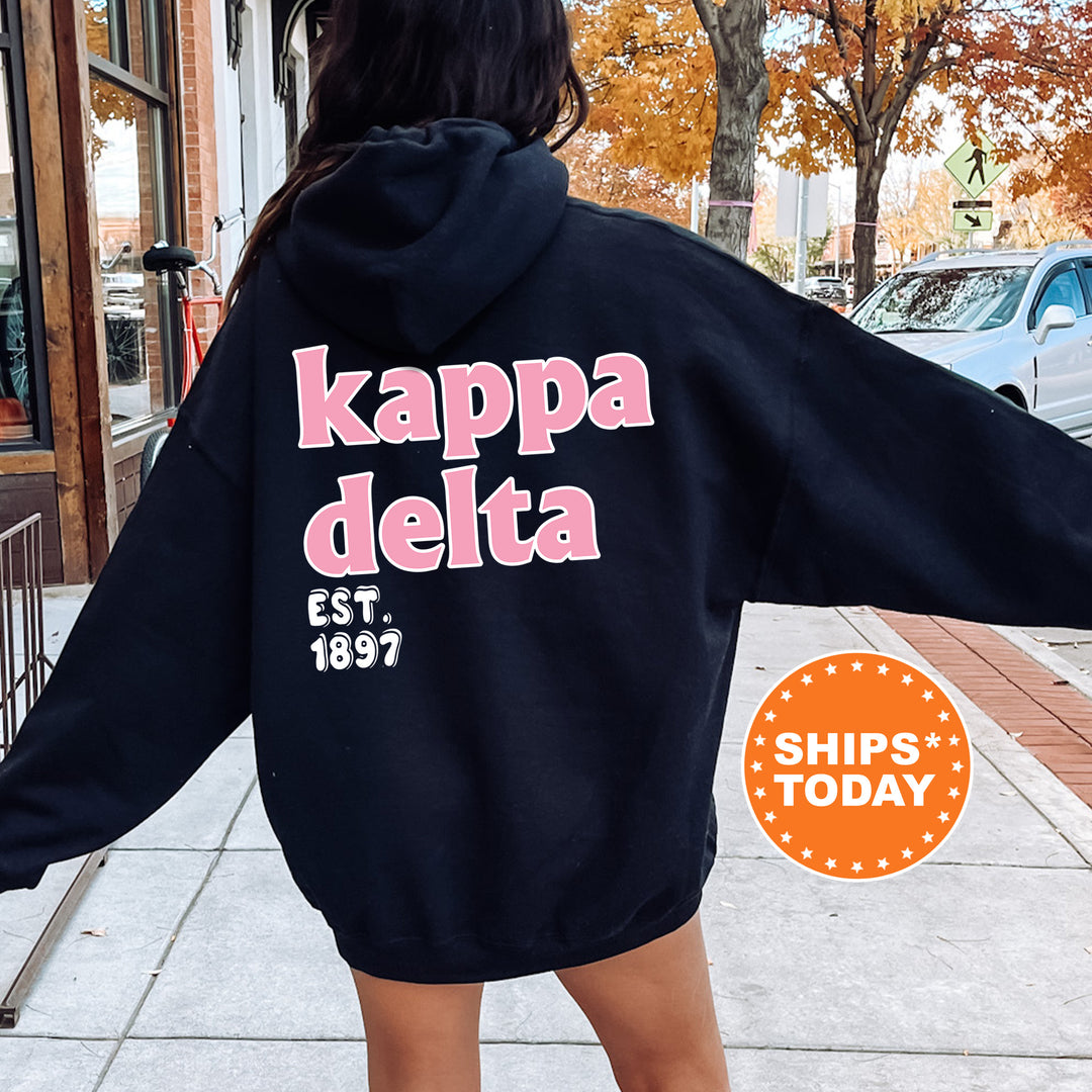 A person wearing a black hoodie with the text " Kappa Delta EST. 1897" printed on it, standing on a sidewalk with a car and a building in the background.